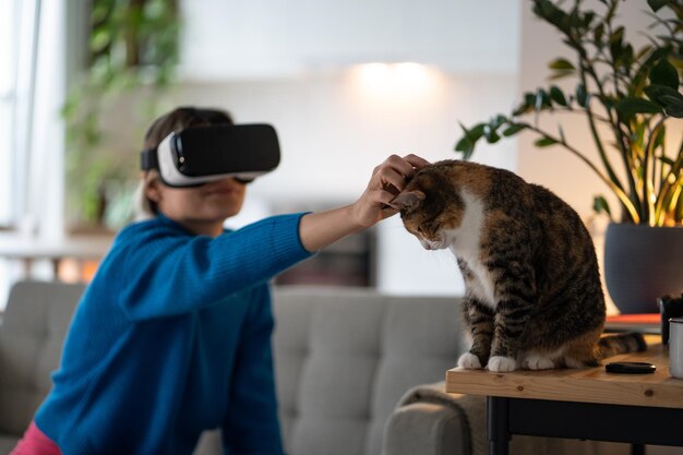 Une femme positive dans un casque VR, un chat caressant, est assise sur un canapé en regardant un film avec des effets de réalité virtuelle