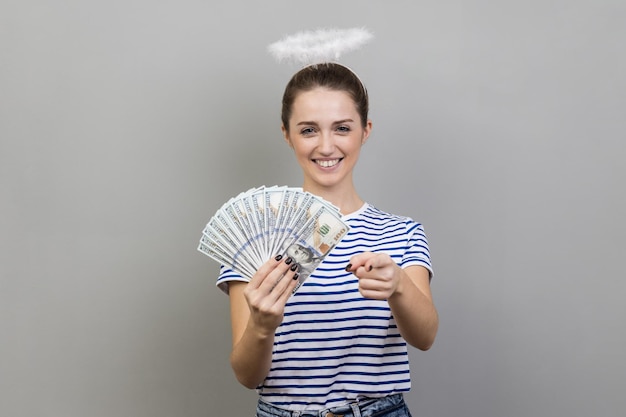 Femme positive avec nimb au-dessus de la tête tenant beaucoup de billets en dollars et pointant vers la caméra