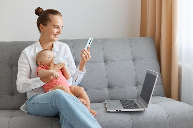 Femme positive souriante portant une chemise blanche et un jean assis sur un canapé avec un bébé dans les mains ayant une expression faciale satisfaite en regardant une vidéo amusante sur un téléphone intelligent