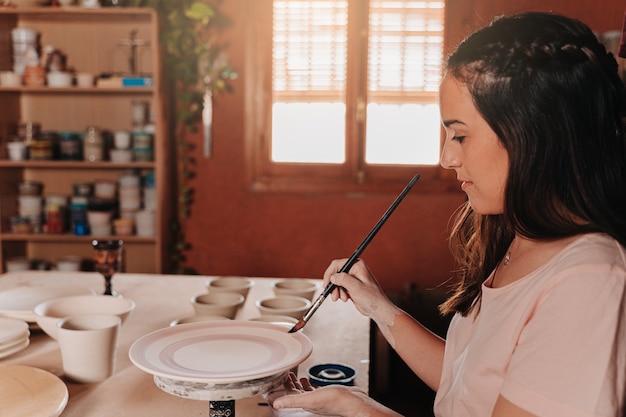 Femme potière dans son atelier peignant ses œuvres d'art sur sa table avec différents outils et couleurs assorties