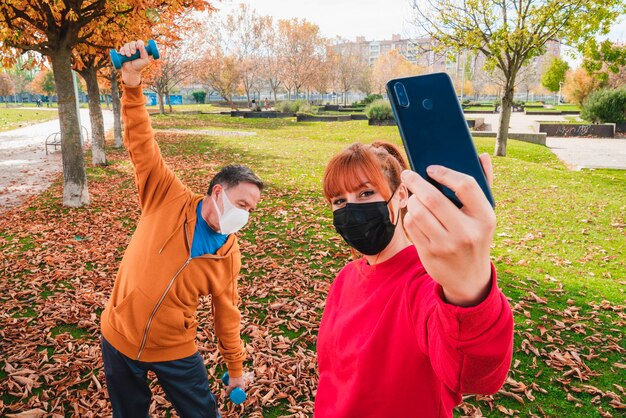 Femme prenant un selfie avec un homme adulte exerçant avec des haltères portant des masques dans un parc