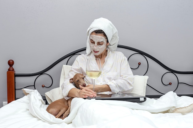 Femme prenant soin de son visage avec un masque au lit, avec un chien, buvant un verre de vin