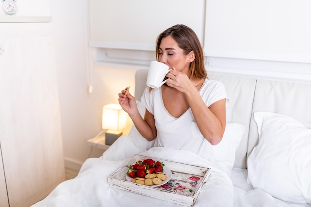 Femme prenant son petit déjeuner et une tasse de café dans la chambre d'hôtel. Plateau avec petit déjeuner sur un lit pour belle belle fille. Belle femme portant et appréciant, petit déjeuner au lit