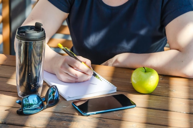 une femme prend des notes dans un cahier pendant le déjeuner, assise sur une véranda par une journée ensoleillée. Apple et café en gobelet sur table