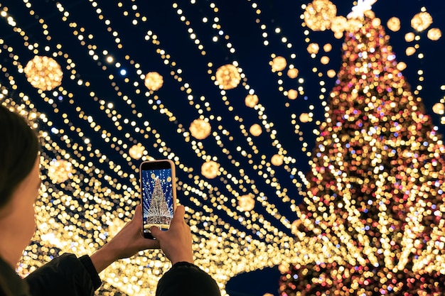 Une femme prend une photo d'un grand arbre de Noël festif urbain sur un smartphone
