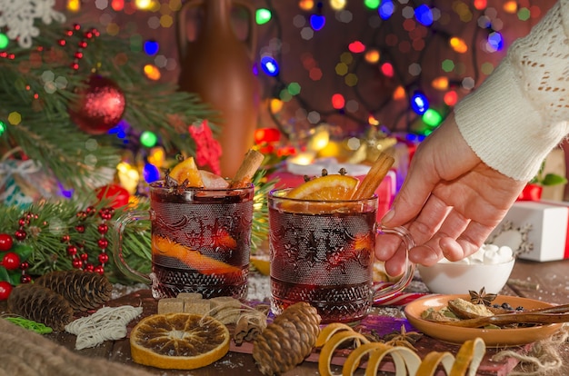 Une femme prend une tasse de vin chaud avec une tranche d'orange sur fond de guirlandes de fête. Style sombre