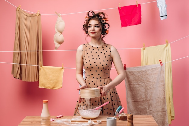 La Femme Prépare La Nourriture, Elle Tamise La Farine à Travers Un Tamis. Belle Femme Au Foyer Est Occupée Aux Tâches Ménagères Sur Fond Rose.