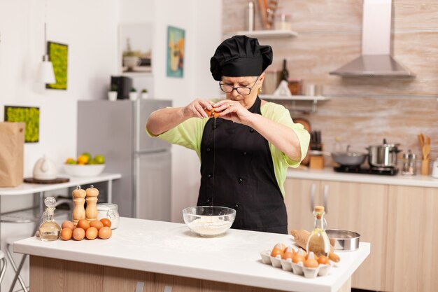 Une femme prépare une pâte pour faire cuire des œufs cassés dans la cuisine à domicile. Pâtissier âgé casser l'oeuf sur un bol en verre pour une recette de gâteau dans la cuisine, mélanger à la main, pétrir les ingrédients en préparant des c maison