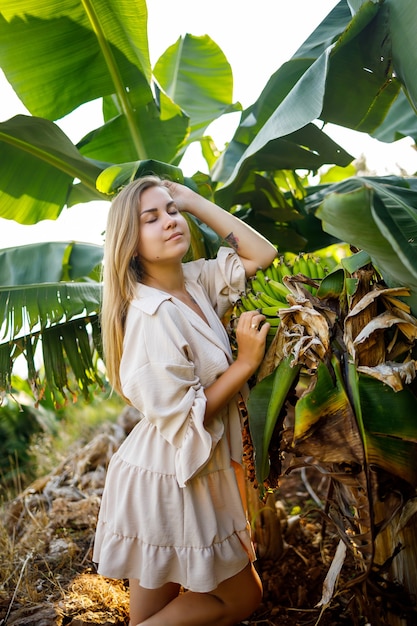 Femme près des feuilles vertes des bananiers sur la nature dans un parc dans un endroit tropical, elle est vêtue d'une jupe beige et d'un chemisier