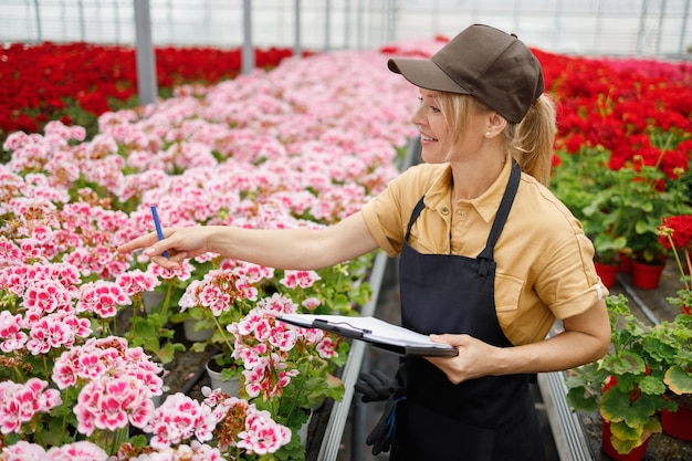 Femme avec presse-papiers inspectant et comptant les plantes dans une serre de fleurs