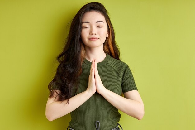 Femme priant avec les mains ensemble pour demander pardon en souriant les yeux fermés, agréable femme brune isolée sur fond vert