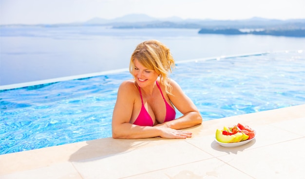 Femme profitant de vacances dans une villa de luxe se relaxant dans une piscine à débordement à côté d'une assiette de fruits frais