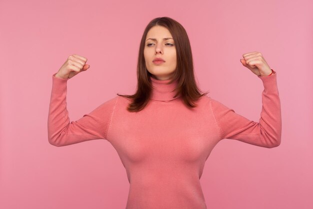 Femme puissante et confiante avec des cheveux bruns levant les mains montrant les muscles des bras démontrant sa force et sa liberté. Studio intérieur tourné isolé sur fond rose