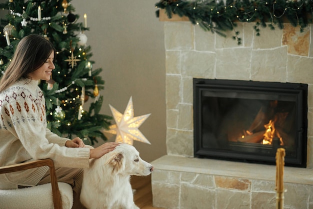 Femme en pull confortable caressant un joli chien blanc sur fond d'arbre de Noël élégant et de cheminée chaleureuse Vacances d'hiver Propriétaire avec animal de compagnie se relaxant dans la salle de fête Joyeux Noël