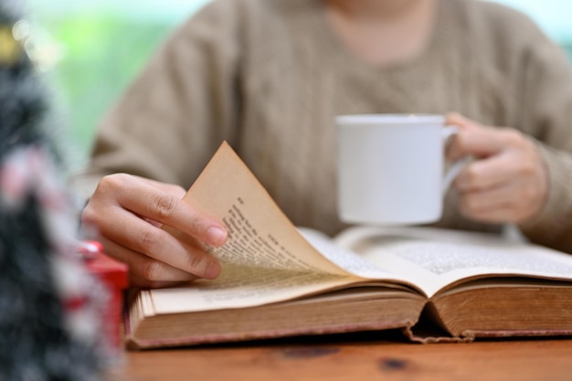 Femme en pull confortable lisant un livre ou un roman tout en sirotant un café chaud à son bureau recadrée