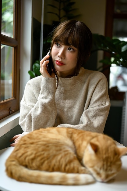 Une femme en pull confortable parlant au téléphone pendant que son chat dort sur une table