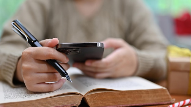 Une femme en pull confortable utilisant son smartphone pour rechercher des informations en ligne tout en lisant un livre