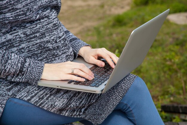 Femme en pull gris et jean bleu tenant un ordinateur portable gris sur ses genoux et l'utilisant