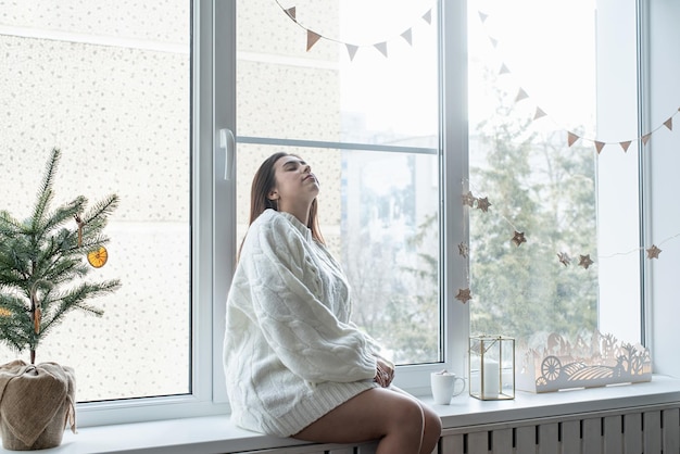 Femme en pull d'hiver blanc chaud assis sur le rebord de la fenêtre à la maison à la veille de noël tenant une tasse avec des guimauves