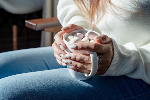 Femme en pull d'hiver blanc chaud tenant une tasse avec des guimauves