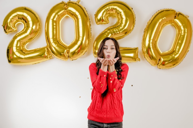 Femme en pull rouge soufflant des confettis argentés devant des ballons du nouvel an 2020