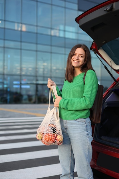 Femme en pull vert tenir un sac à provisions avec une épicerie près du coffre des voitures rouges