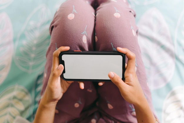 Photo une femme en pyjama est allongée sur un lit et regarde un smartphone avec un écran blanc.