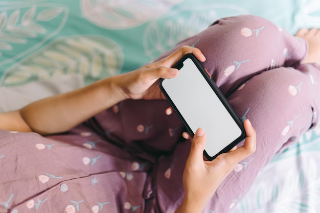 Photo une femme en pyjama est allongée sur un lit et regarde un smartphone avec un écran blanc.