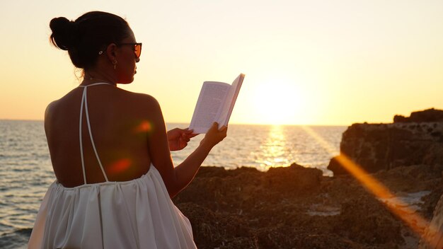 Une femme qui lit au coucher du soleil sur la côte rocheuse aime un livre