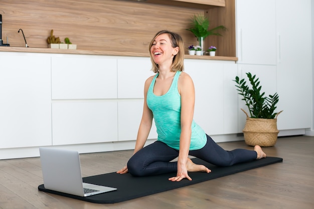 Femme qui rit qui s'étend devant l'ordinateur portable tout en faisant du yoga en ligne à la maison