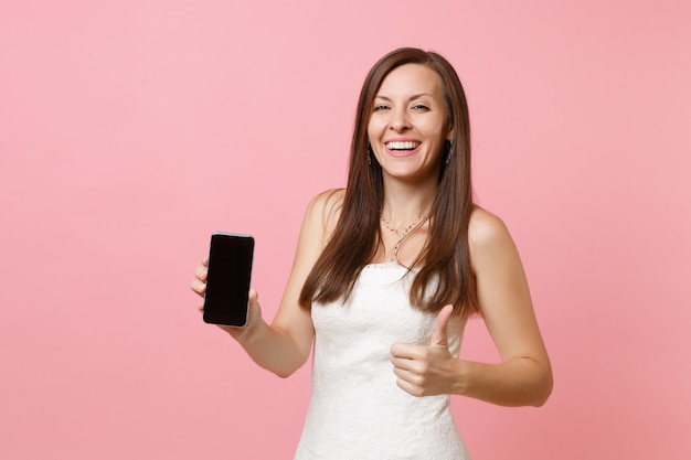 Femme qui rit en robe blanche montrant le pouce vers le haut, tenir un téléphone portable avec un écran vide noir vierge