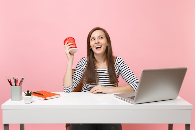 Femme qui rit tenant une tasse de café ou de thé travaillant sur un projet assis au bureau avec un ordinateur portable isolé sur fond rose pastel. Concept de carrière d'entreprise de réalisation. Copiez l'espace pour la publicité.