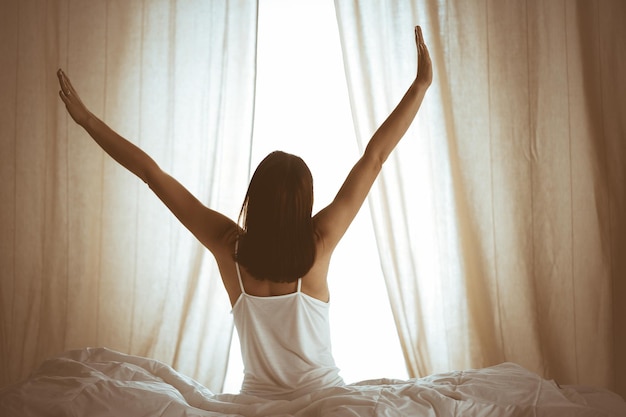 Femme qui s'étire les mains dans son lit après le réveil, éruption solaire. Brunette entrant dans une journée heureuse et détendue après une bonne nuit de sommeil et une vue arrière. Concept d'un nouveau jour et d'un week-end joyeux.