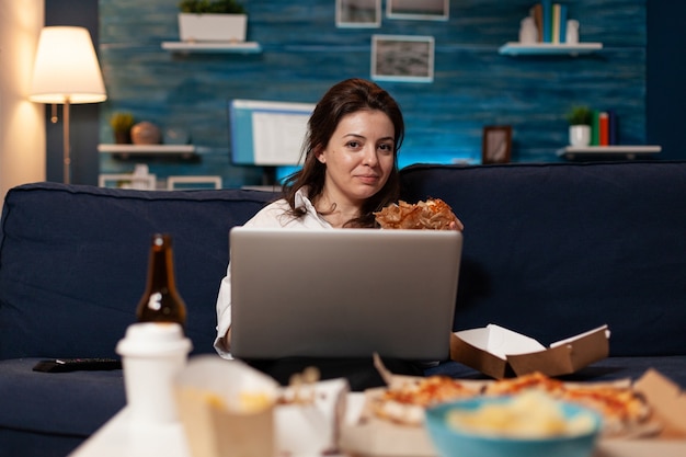 Femme de race blanche assise sur un canapé en train de manger un délicieux hamburger délicieux tout en travaillant sur un ordinateur portable