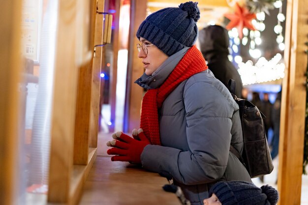 Femme de race blanche portant un chapeau et une écharpe rouge achetant des friandises en kiosque au marché de Noël. Imahe avec mise au point sélective et bruit