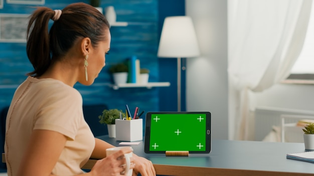 Femme De Race Blanche Regardant Une Tablette Avec Une Clé De Chrominance D'écran Vert Maquette Assise Sur Un Bureau Dans Le Salon. Femme Indépendante Naviguant Sur Un Réseau Social à L'aide D'un Appareil Isolé