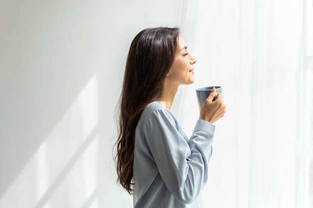 une femme de race blanche regarde le paysage par la fenêtre et profite du premier café du matin au soleil