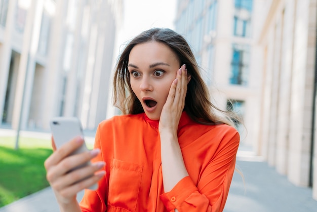 Une femme de race blanche se demande et est choquée en regardant un smartphone dans ses mains.