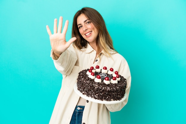 Femme de race blanche tenant un gâteau d'anniversaire isolé sur fond bleu comptant cinq avec les doigts