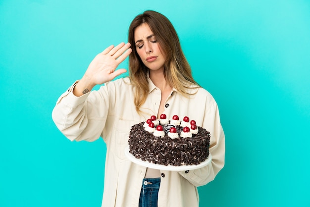 Femme de race blanche tenant un gâteau d'anniversaire isolé sur fond bleu faisant un geste d'arrêt et déçu