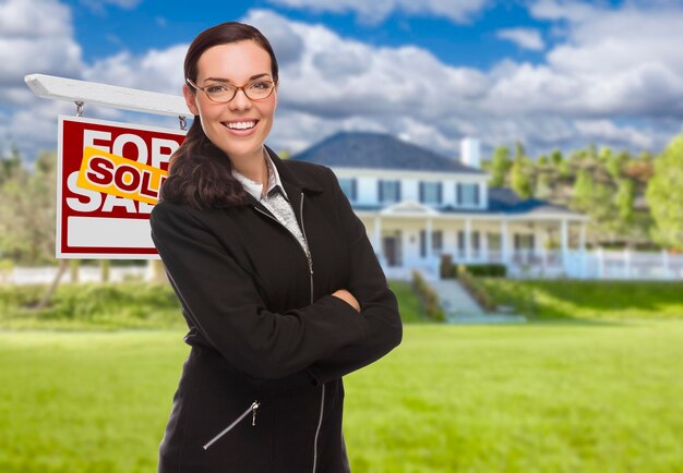 Une femme de race mixte devant une maison et un panneau de vente