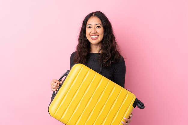 Femme de race mixte sur mur rose isolé en vacances avec valise de voyage et un chapeau