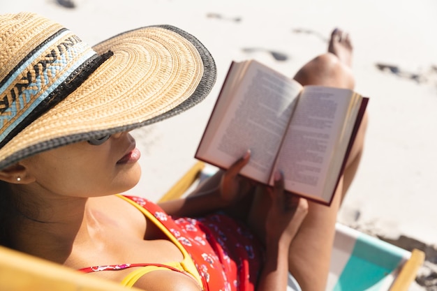 Femme de race mixte en vacances à la plage assis dans un transat lisant un livre. loisirs en plein air sains au bord de la mer.