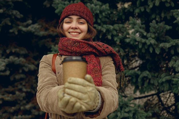 Femme ravie en chapeau chaud et manteau montrant une tasse de café à emporter et regardant la caméra