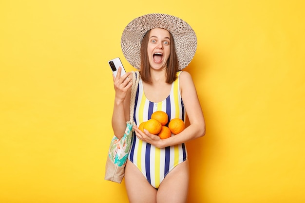 Une femme ravie portant un maillot de bain isolé sur fond jaune tenant des fruits et un smartphone reçoit des sms sur le départ pour les vacances en réservant des billets en ligne