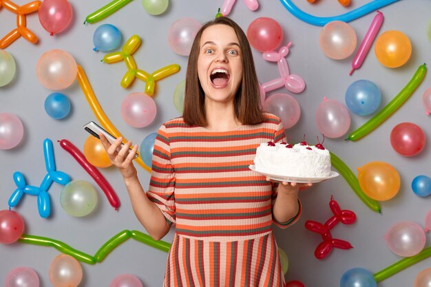 Femme ravie positive aux cheveux bruns portant une robe rayée tenant un gâteau et un téléphone intelligent dans les mains criant d'excitation debout contre un mur gris décoré de ballons colorés