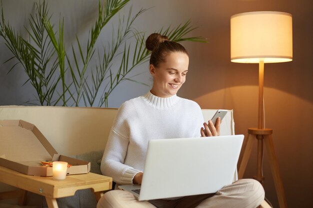 Femme ravie souriante avec une coiffure chignon portant un pull blanc assis sur un canapé et travaillant sur un ordinateur portable tenant un téléphone portable dans les mains et vérifiant les e-mails ou les réseaux sociaux
