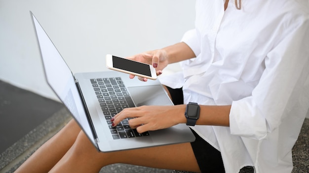 Femme recadrée en chemise blanche assise dans les escaliers à l'aide d'un smartphone et tapant sur un clavier d'ordinateur portable