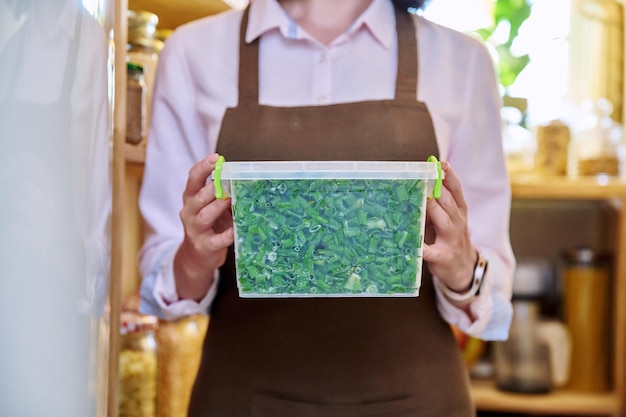 Femme avec récipient d'oignons verts congelés dans la cuisine dans le garde-manger près du congélateur