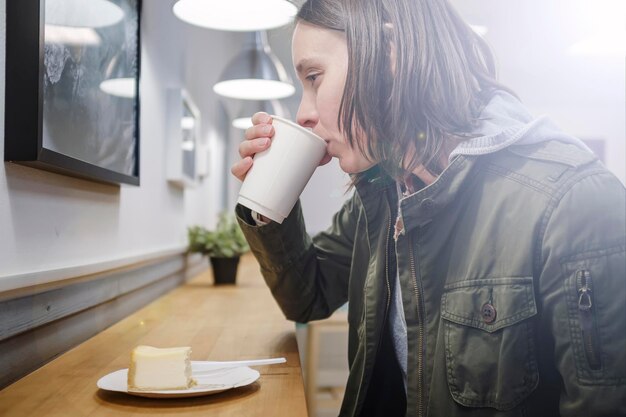 Femme réfléchie buvant du cappuccino dans une tasse en papier et mangeant un gâteau au fromage dans un café confortable